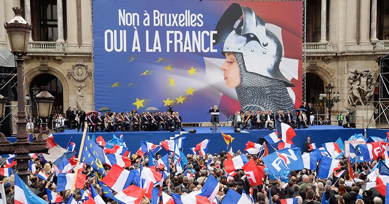 Supporters of France's far-right National Front (FN) wave French flags as leader Marine Le Pen (rear C) delivers a speech in front of a poster depicting Joan of Arc (Jeanne d'Arc) and reading "No to Brussels, yes to France" during a May Day rally in Paris on May 1, 2014. AFP PHOTO / PIERRE ANDRIEU (Photo credit should read PIERRE ANDRIEU/AFP/Getty Images)